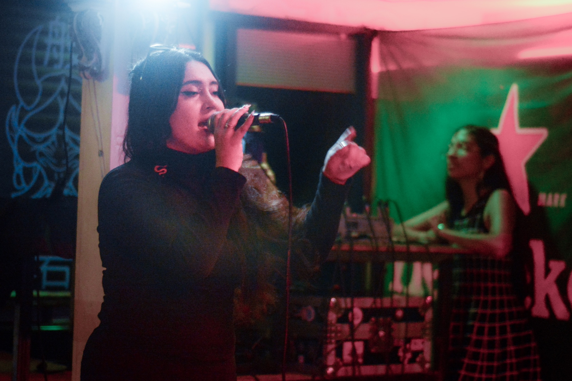 Cantante con micrófono y productora al fondo bajo luces verdes y rojas