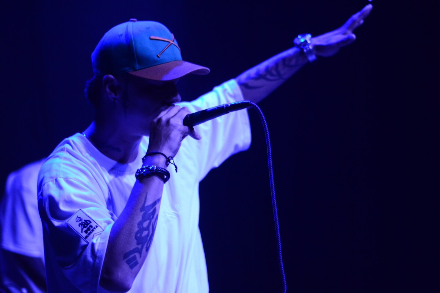 MC con gorra y micrófono bajo luz azul, brazo extendido